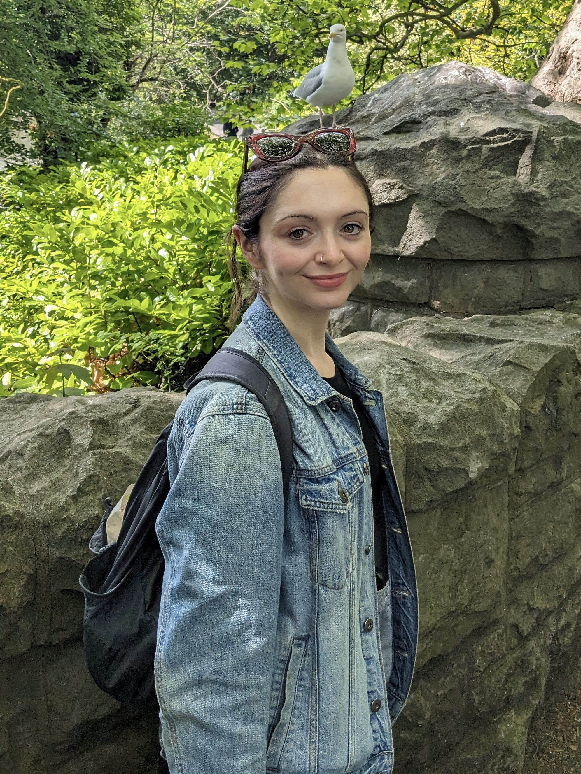 white feminine looking person standing outside. a stone wall is behind her. she is wearing a denim jacket. she is smiling.